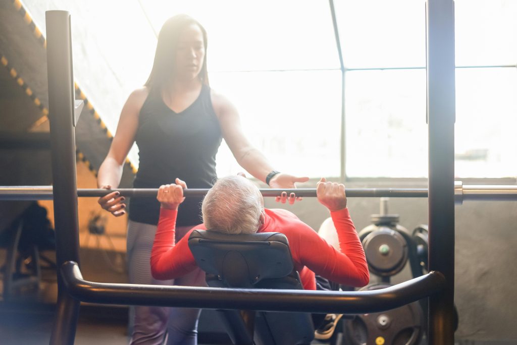 Elderly man in gym performing bench press with female trainer assistance, promoting fitness and health.